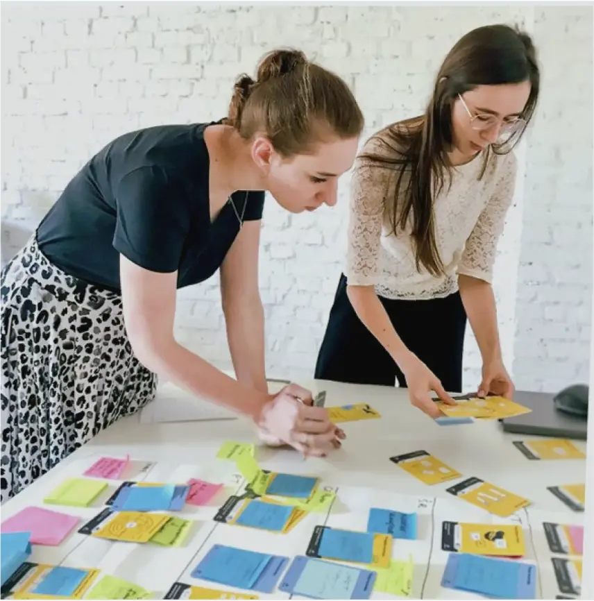 Two young women are leaning intently over a table, planning strategic workshops. They use Witflow cards and colorful sticky notes to plan the workshops.