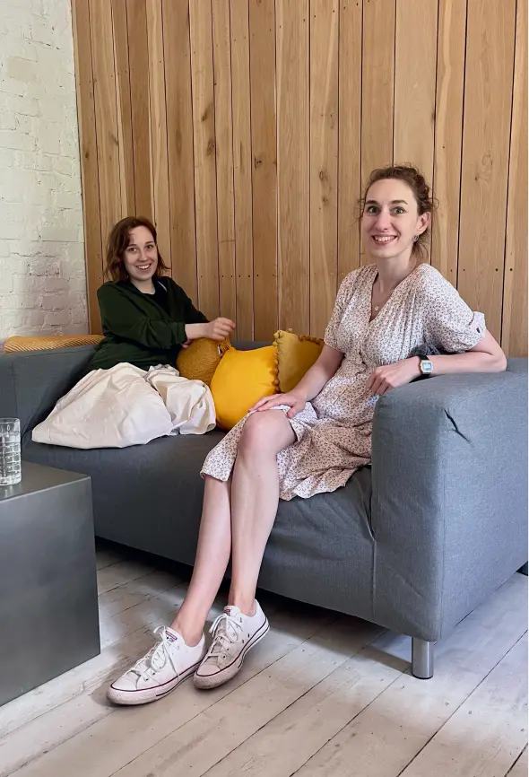 Two women dressed in summer clothes are sitting on a gray sofa with yellow pillows. They look straight into the camera with big smiles and positive attitudes.