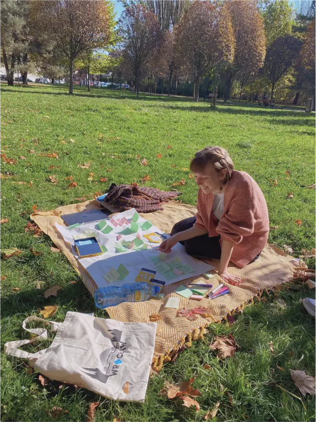 A smiling woman in a pink sweater sits on a blanket in a park with green grass. She has a workshop planning matrix spread out in front of her and a coloured marker in her hand.
