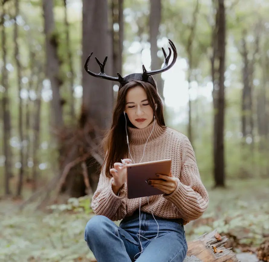 A young woman sits on a stump in the forest and draws on a tablet. She is dressed in a cream sweater and jeans, has antlers on her head, and headphones in her ears.