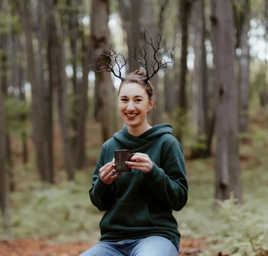 A smiling woman sitting in the forest looks straight at the camera. She is dressed in a green sweatshirt, holds a cup of coffee in her hands, and has antlers on her head.
